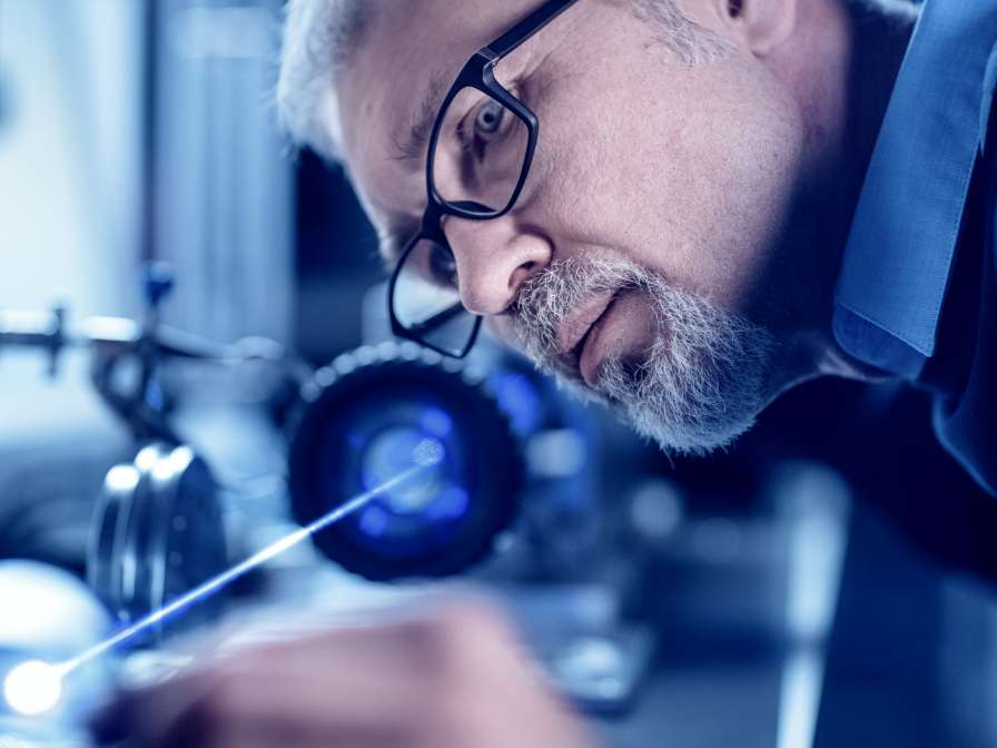 Close-up Portrait of Focused Middle Aged Engineer in Glasses Working with High Precision Laser Equipment, Using Lenses and Testing Optics for Accuracy Required Electronics